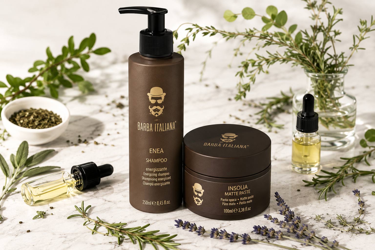 Натуральна чоловіча косметика Barba Italiana, представлена шампунем ENEA та матовою пастою INSOLIA на білій кам'яній поверхні в оточенні свіжих трав та ефірних олій.