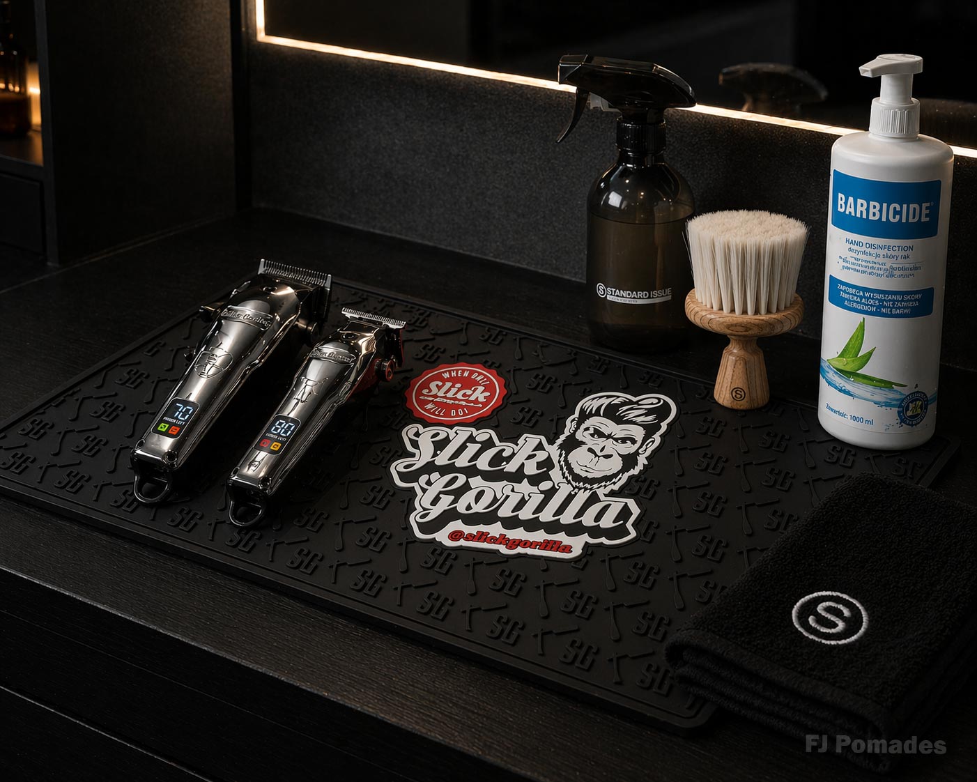 Робоче місце барбера з чорним силіконовим килимком Slick Gorilla, на якому розташовані два сріблясті професійні тримери з цифровими дисплеями, чорний пульверизатор, дерев’яна щітка для фейдингу, флакон антисептика для рук Barbicide та згорнутий чорний рушник із логотипом.
